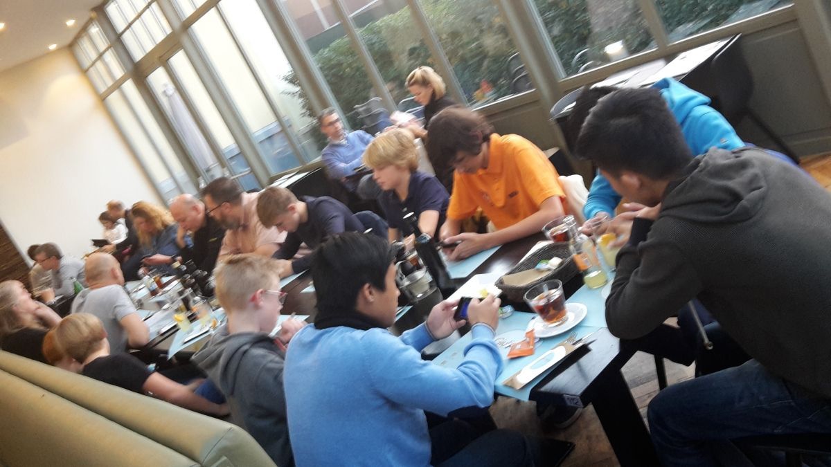 Mensen zaten aan tafeltjes in een restaurant, sommigen telefonerend, anderen pratend. Natuurlijk licht, neutrale tinten.