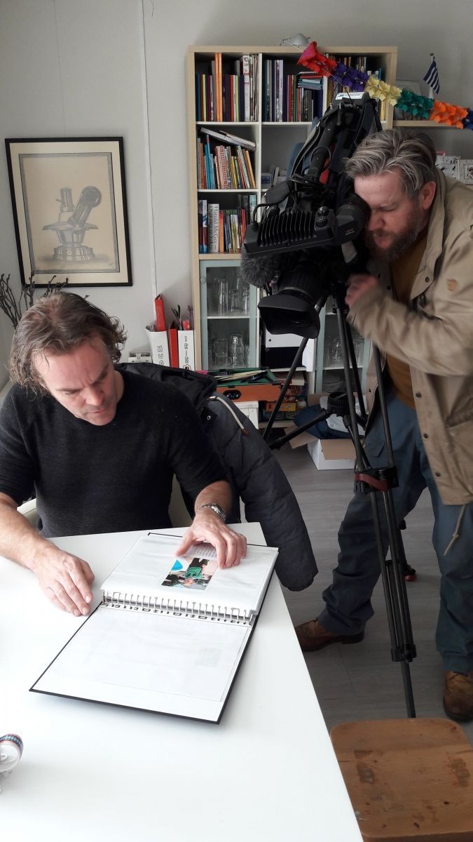 Man gefilmd door een cameraman. Man in het zwart kijkt in een map, de cameraman kijkt door de camera.