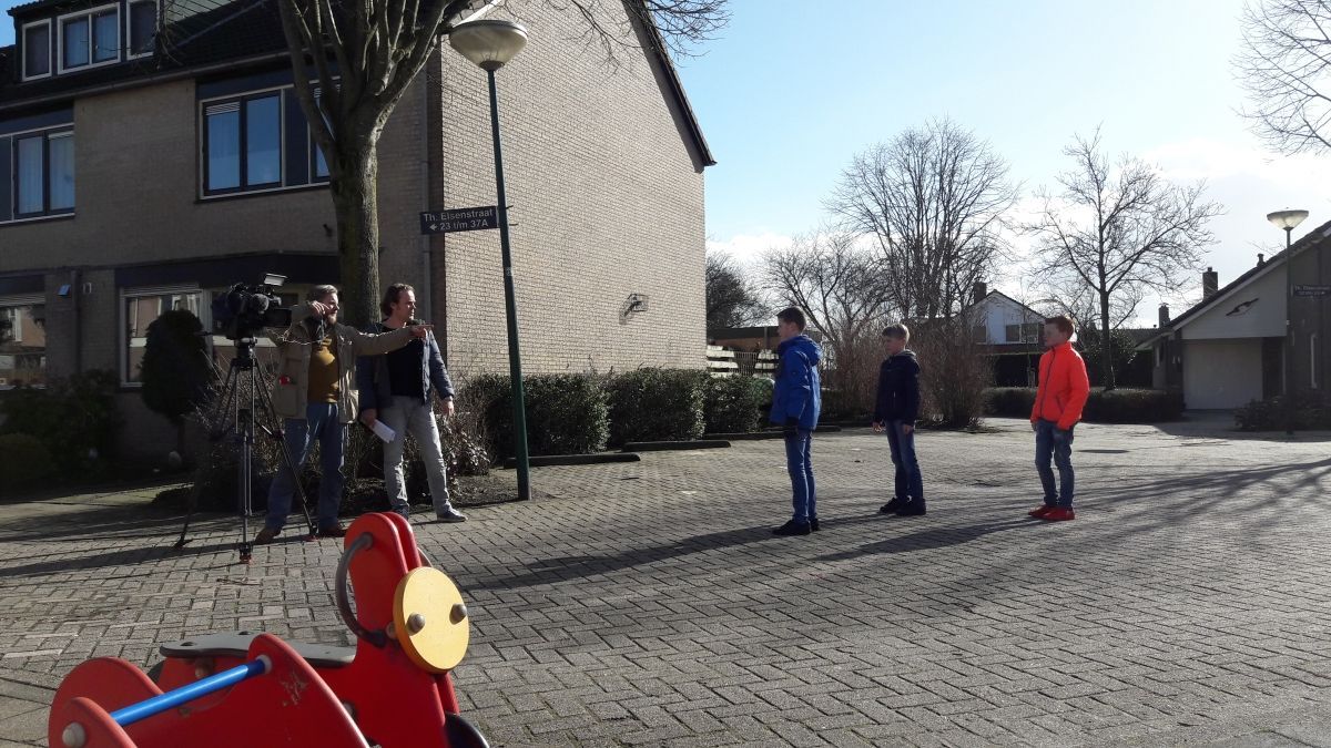 Kinderen in kleurrijke jasjes staan ​​op een rij terwijl een filmploeg hen voor een huis filmt.