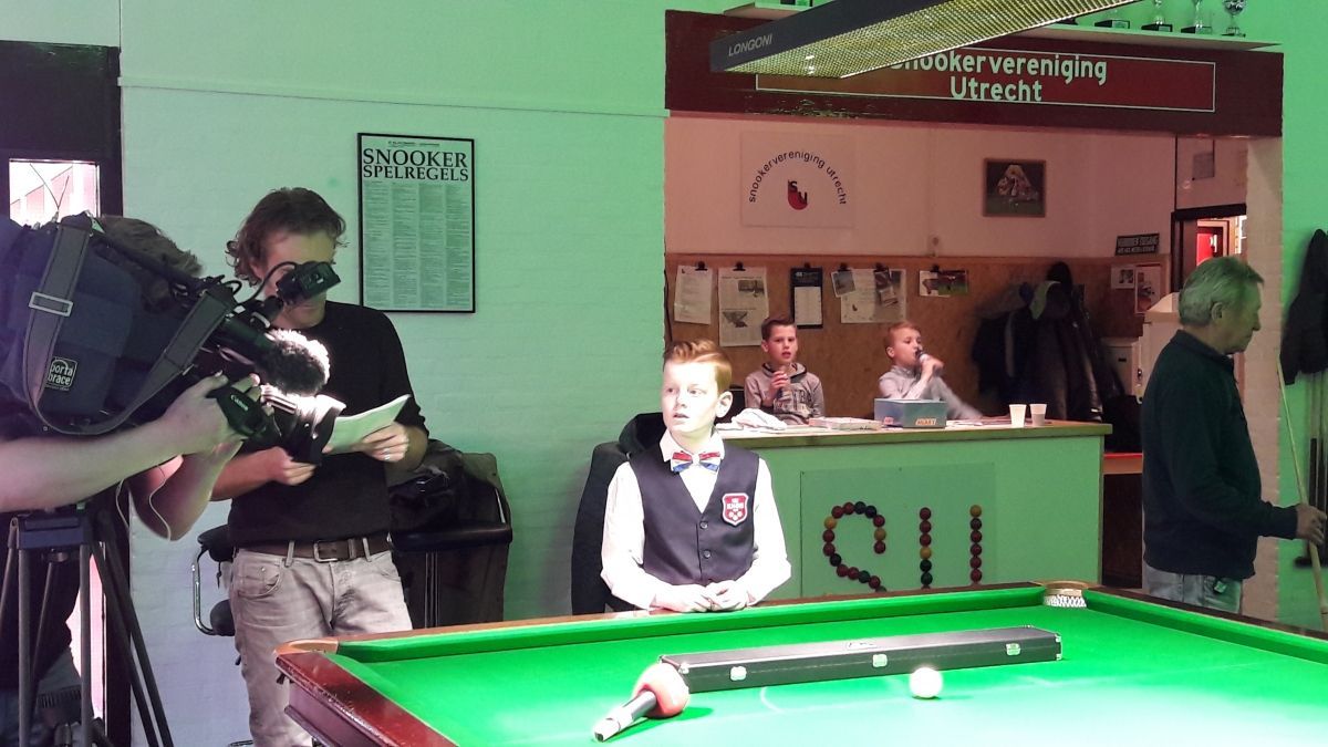 Man filmt snookerwedstrijd; jonge speler aan tafel, anderen op de achtergrond bij de scorebalie.