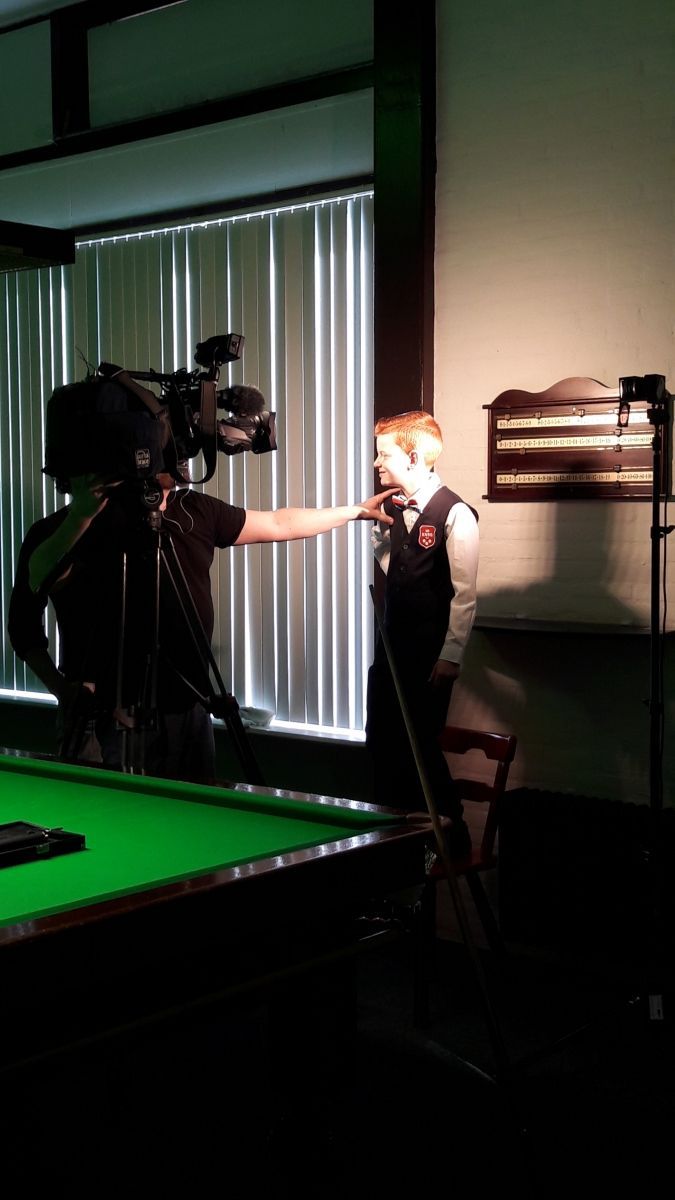 Een persoon in een vest wordt geïnterviewd bij een pooltafel. Een cameraman richt een camera op hem in een schemerige kamer.