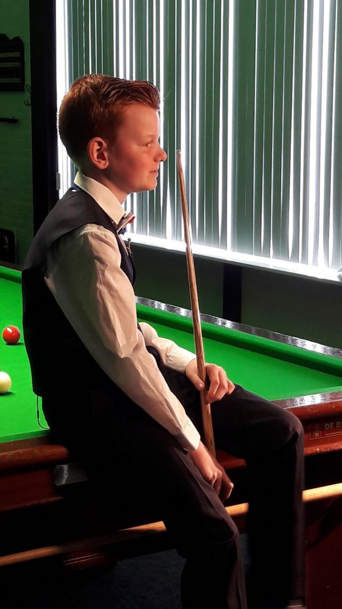 Jongeman in formele kleding zit op een snookertafel, houdt een keu vast en de ballen liggen zichtbaar op de tafel.