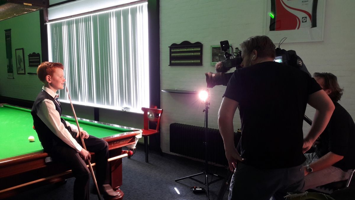 Een persoon in formele kleding zit op een pooltafel en wordt gefilmd door een crew in een snookerhal.