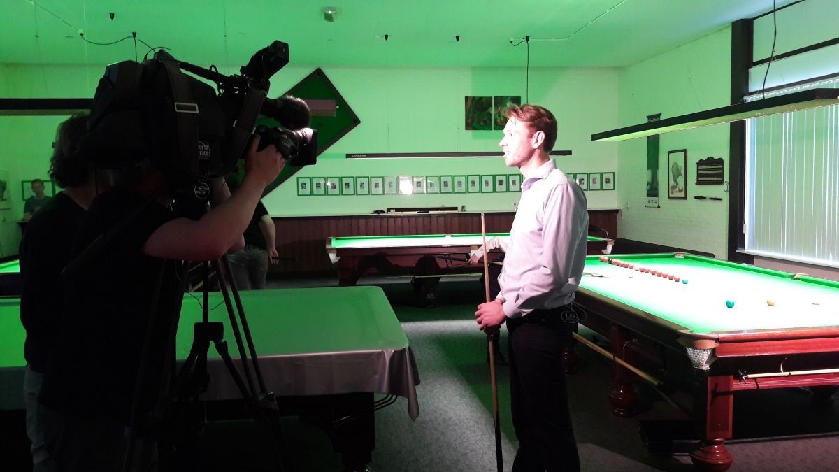 Een man met een snookerkeu wordt gefilmd in een snookerhal. Camera en crew op de voorgrond.