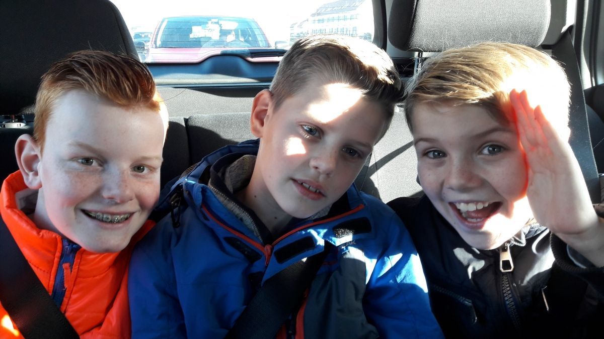 Drie jongens lachen en zwaaien vanaf de achterbank van een auto. Eén draagt ​​een bretels en een oranje jasje.