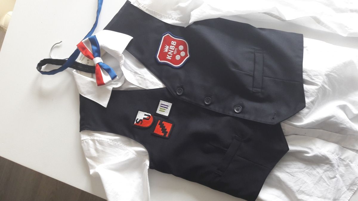 Marineblauw vest met patches over een wit overhemd, met een rood-wit-blauwe vlinderdas aan een hanger.