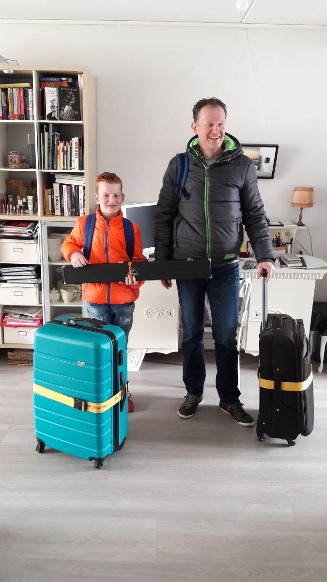 Vader en zoon klaar voor vertrek, lachend. Jongen met blauwe koffer, vader met zwarte. Binnen in een huis zijn de tassen vastgebonden.