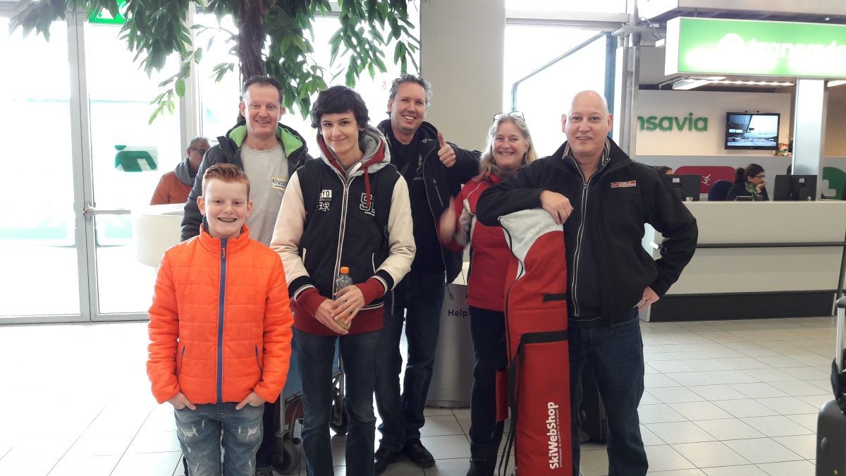 Een groep van zeven glimlachende mensen poseren binnen. Sommigen dragen rode tassen. Feloranje jasjes, donker haar, luchthavenomgeving.