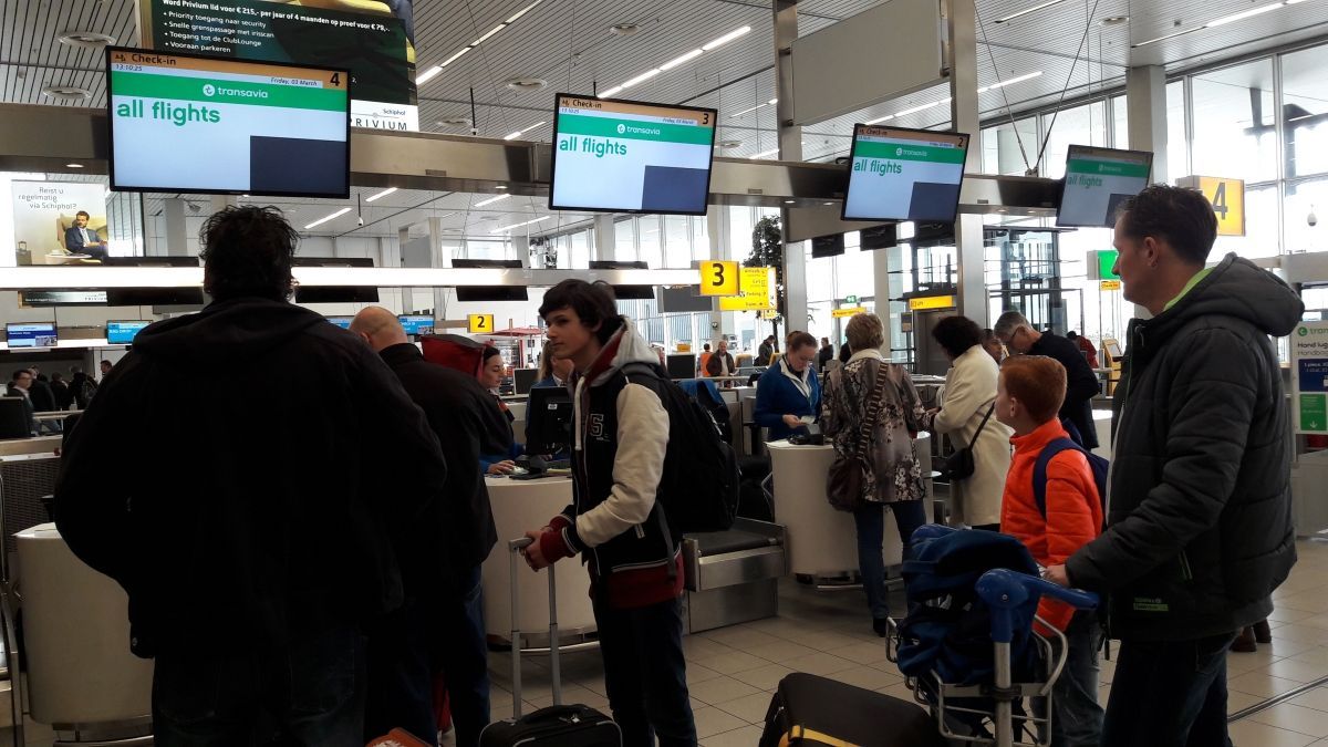 Mensen in de rij bij de incheckbalies op het vliegveld, met schermen waarop vluchtinformatie wordt weergegeven.