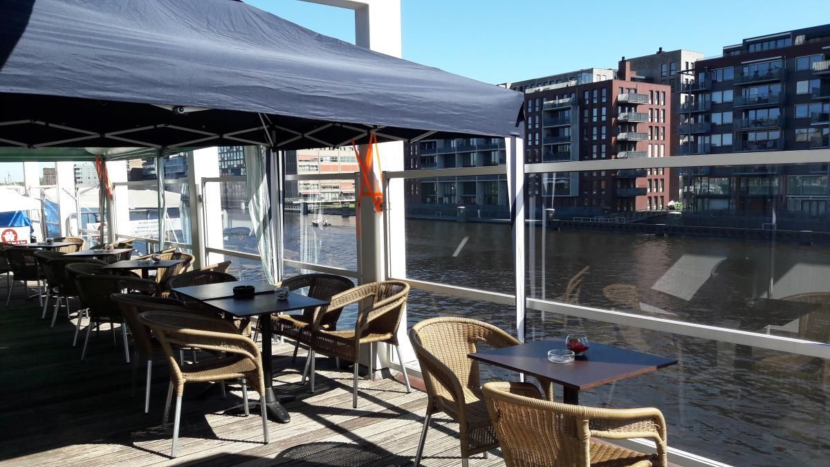 Terras met terras aan de rivier, met tafels, rieten stoelen en een donkerblauwe luifel.
