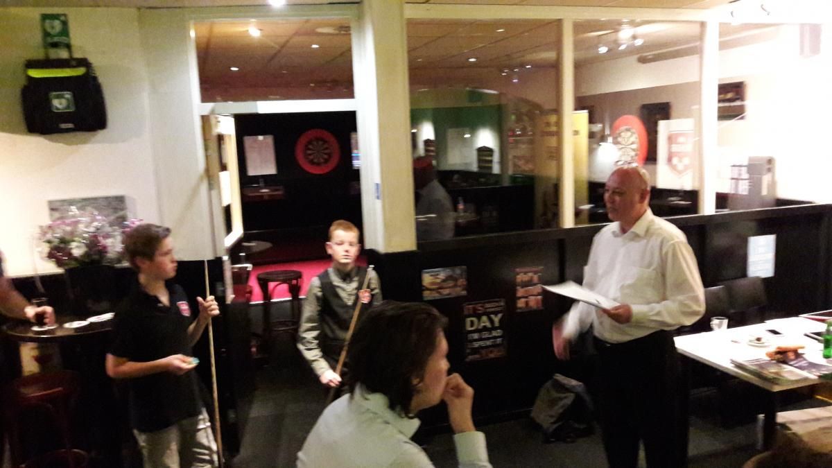 Een man spreekt, twee jongens houden keuen vast en een vrouw zit in een kroeg.