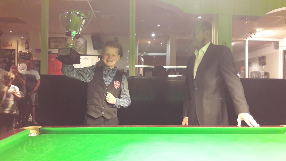 Een jonge snookerkampioen met een trofee in zijn hand, lachend op een groene tafel, met een volwassene in pak naast hem.