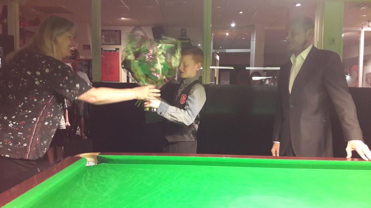 Een vrouw geeft bloemen aan een jonge jongen op een pooltafel. Een man in pak staat ernaast.