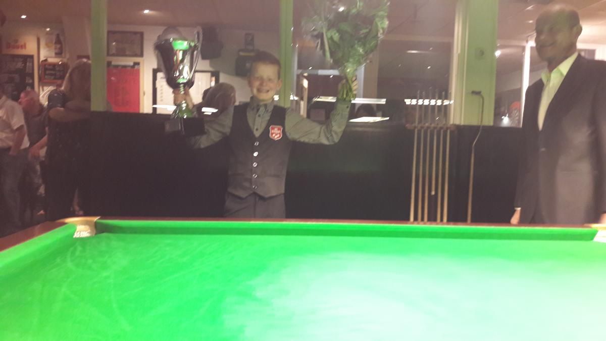 Een snookerspeler steekt een trofee en bloemen omhoog en viert de overwinning aan een pooltafel. Er staat iemand naast hem.