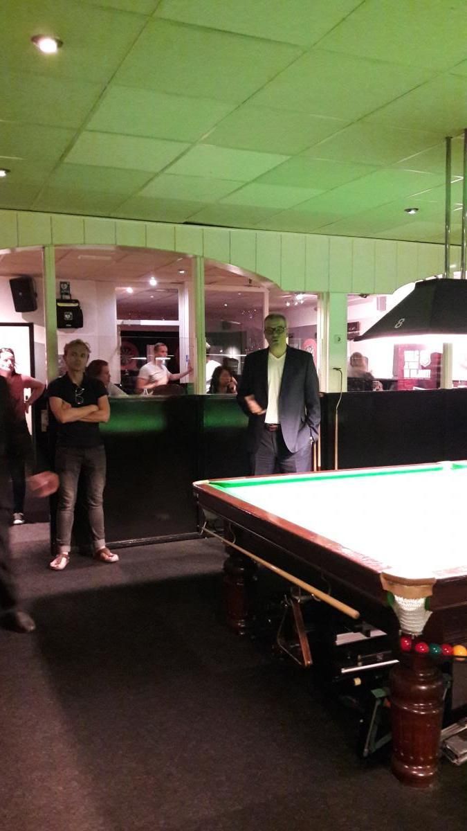 Man in pak staat bij een pooltafel, anderen kijken toe. Groen verlicht interieur.