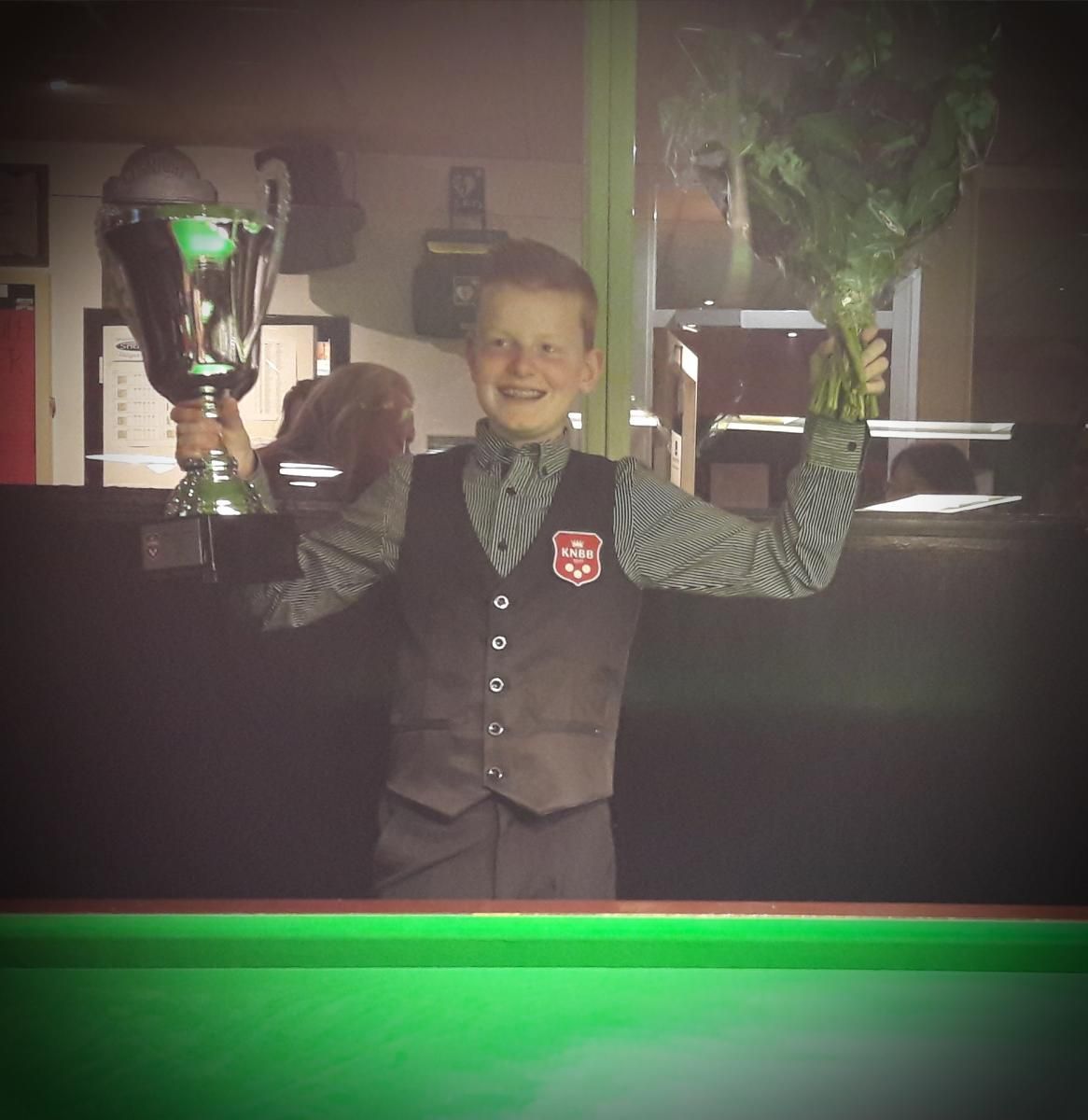 Jongen in vest houdt trofee en bloemen vast en viert de overwinning bij een groene pooltafel.