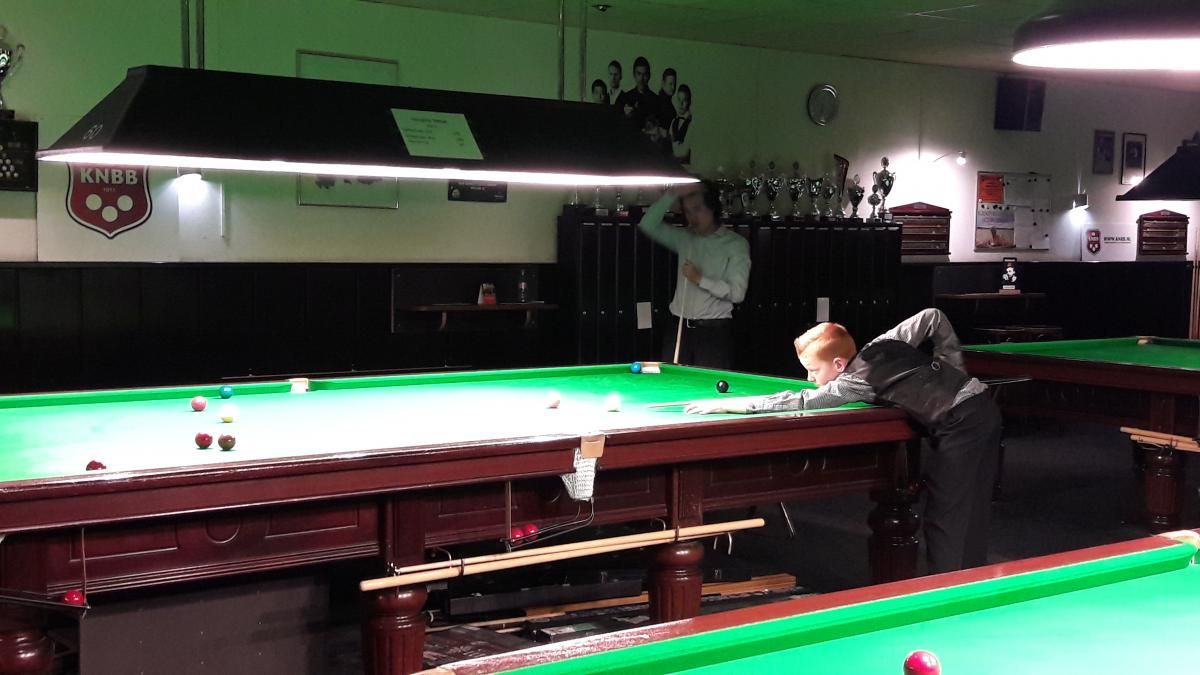 Iemand die snooker speelt in een kamer met weinig licht, terwijl iemand anders toekijkt.