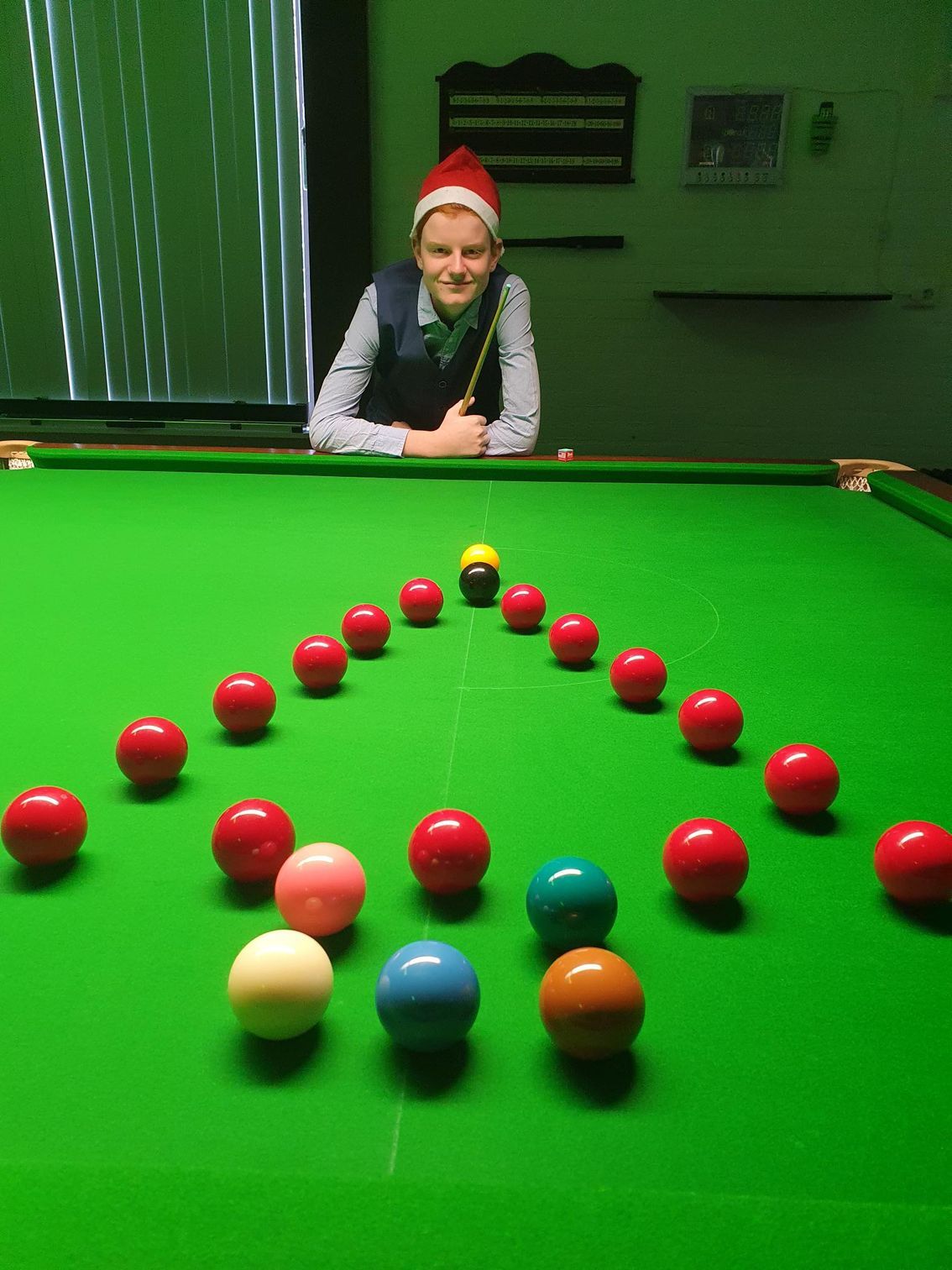 Een persoon met een kerstmanmuts leunt op een snookertafel waarop ballen liggen: groen vilt, rode en gele ballen.