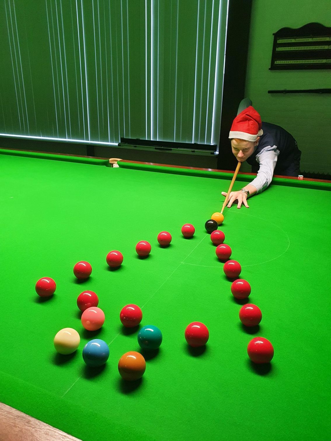 Persoon met Kerstmanmuts speelt snooker, mikkend op een driehoek van rode ballen op een groene tafel.