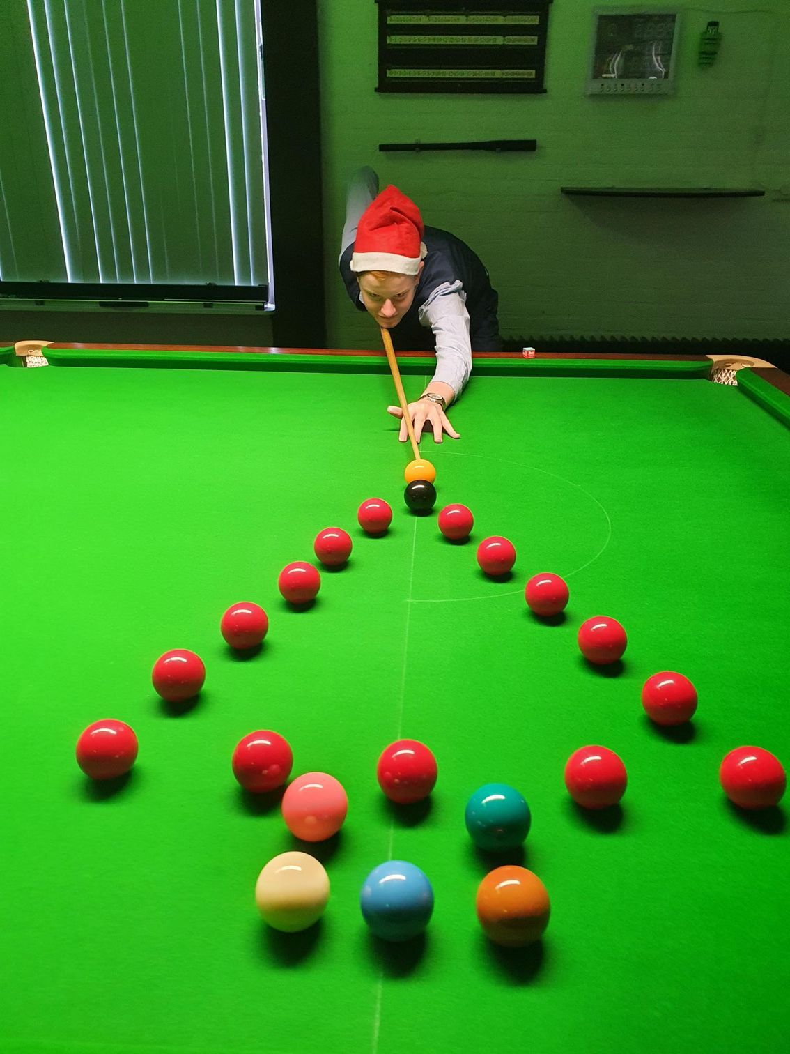 Een persoon met een kerstmanmuts legt een bal op een snookertafel, waarbij de ballen in de vorm van een kerstboom zijn gerangschikt.