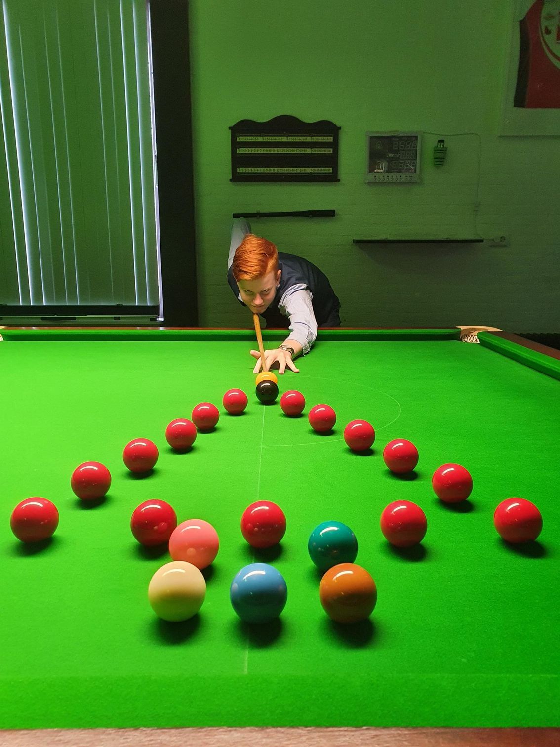 Jongere met rood haar speelt snooker, mikkend op een zwarte bal te midden van een driehoek van rode ballen op een groene tafel.