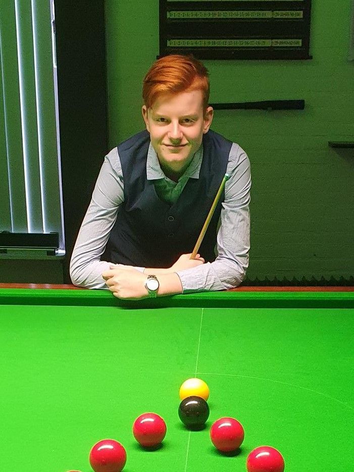 Een jongeman met rood haar leunt op een snookertafel, houdt een keu vast en ziet de ballen liggen.
