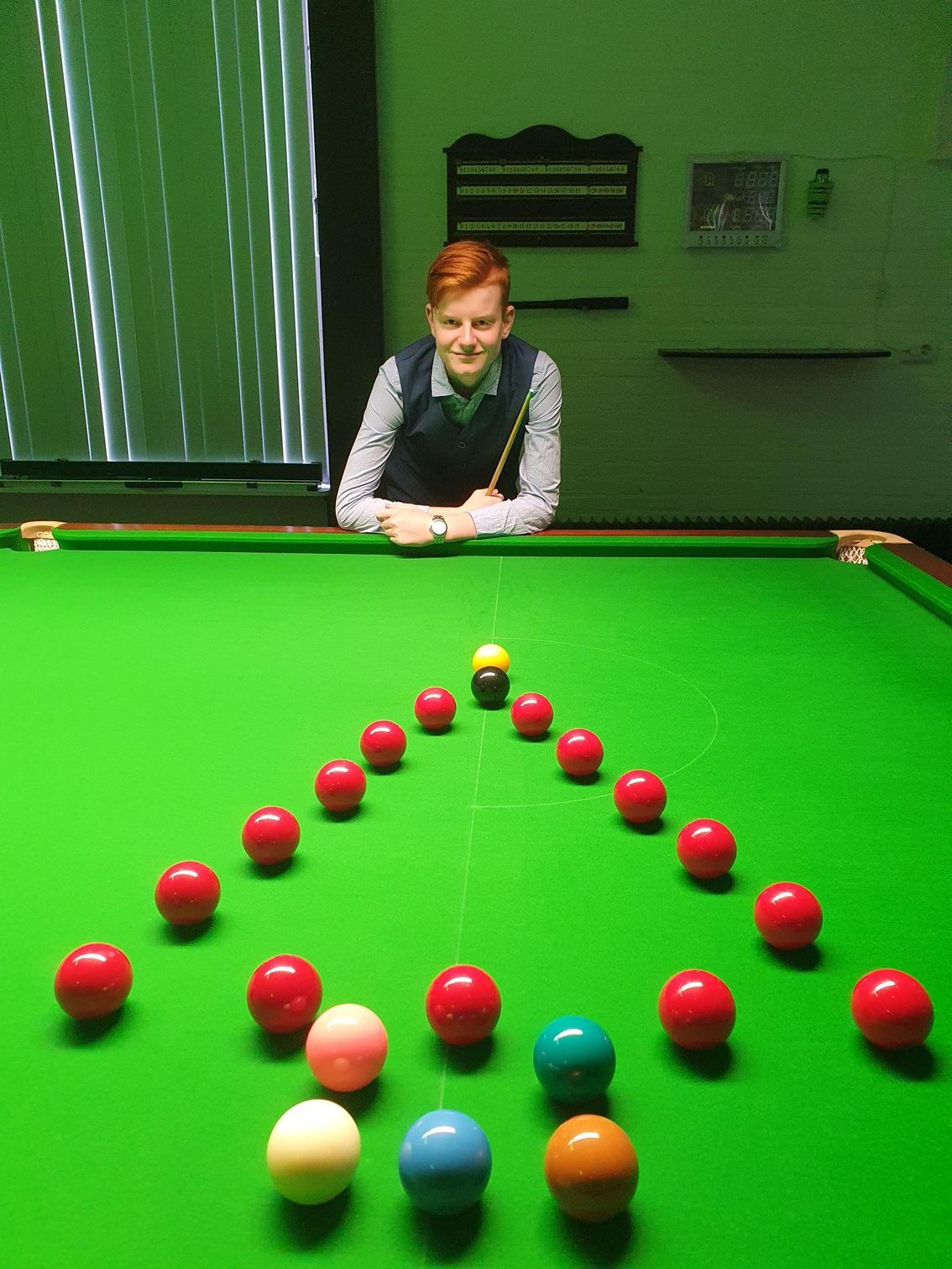 Jongere met keu staat aan een snookertafel; rode ballen in een driehoek, gekleurde ballen op de voorgrond.