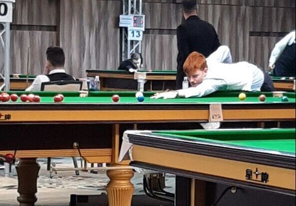 Jongen met rood haar speelt snooker, leunend over de tafel. Andere spelers en tafels in een grote zaal.