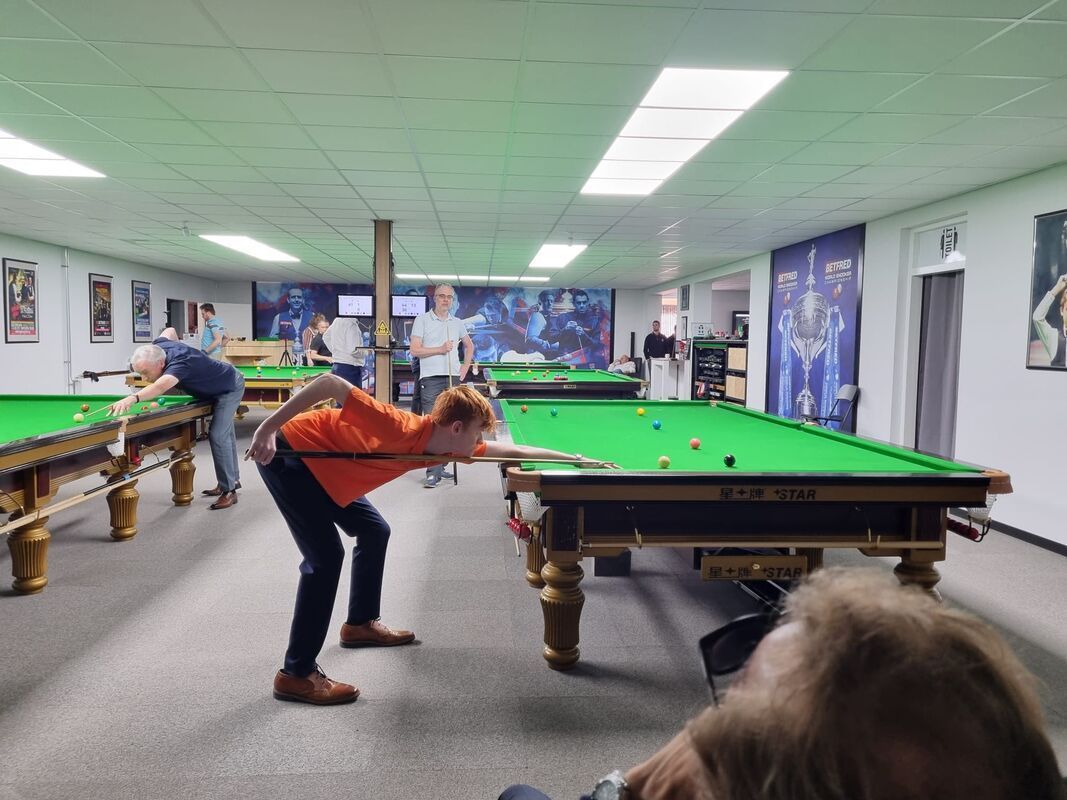 Een persoon in een oranje shirt bereidt zich voor op een potje snooker aan een snookertafel in een snookerhal.