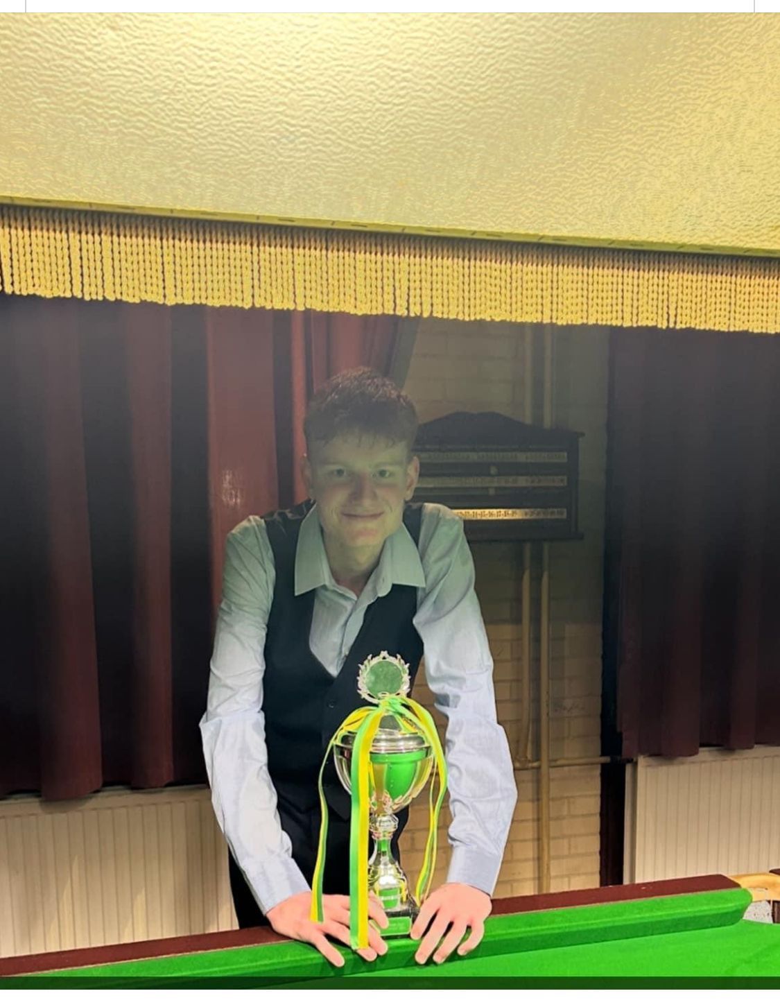 Jongeman in vest, glimlachend, met een trofee op een pooltafel. Groene, gouden trofee. Donkere gordijnen op de achtergrond.