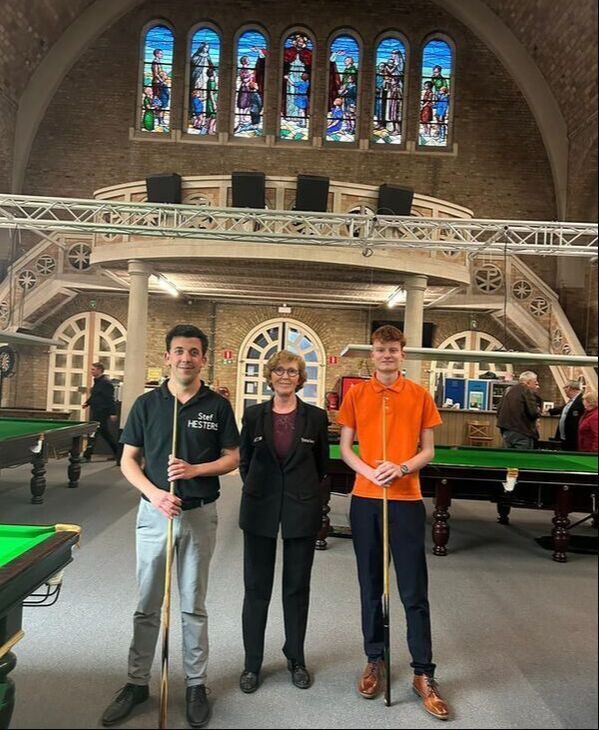 Drie mensen met snookerkeuen poseren in een snookerzaal met glas-in-loodramen.