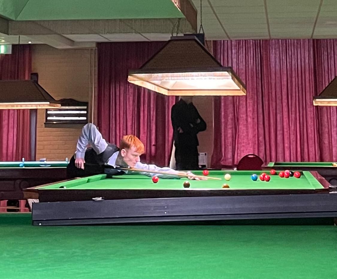 Man in pak speelt snooker op een groene tafel, onder lampen, een andere man op de achtergrond en een rood gordijn.