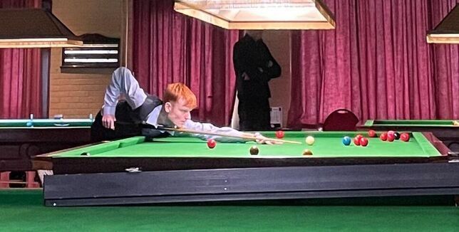 Een snookerspeler met rood haar plaatst een bal op een groene tafel; op de achtergrond staat een toeschouwer.