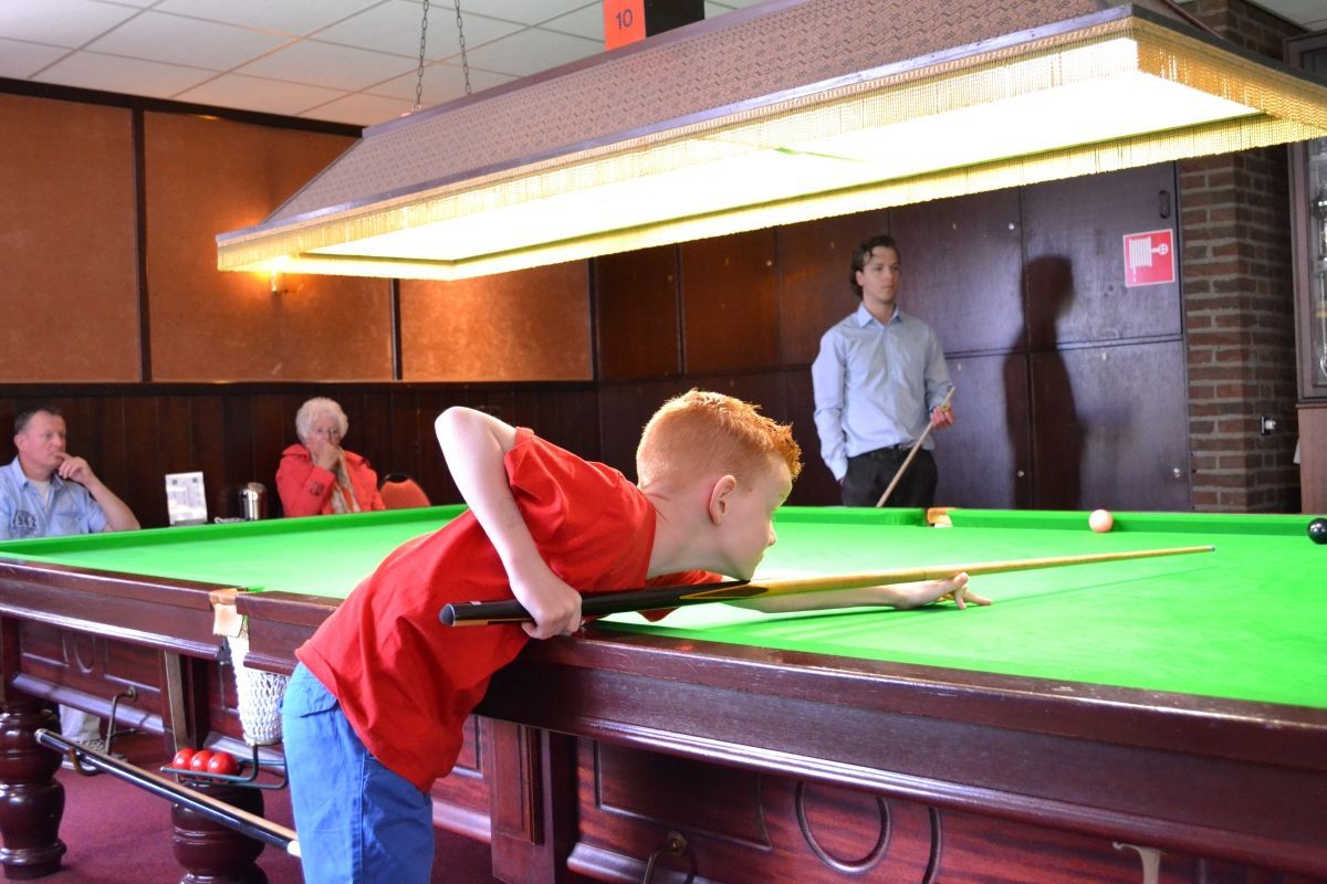 Een jongen bereidt zich voor op een potje pool, onder fel licht. Anderen kijken toe in een café.