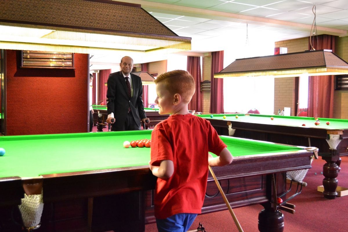 Een jonge jongen in een rood shirt maakt zich klaar om te poolen, terwijl een oudere man in een pak toekijkt.