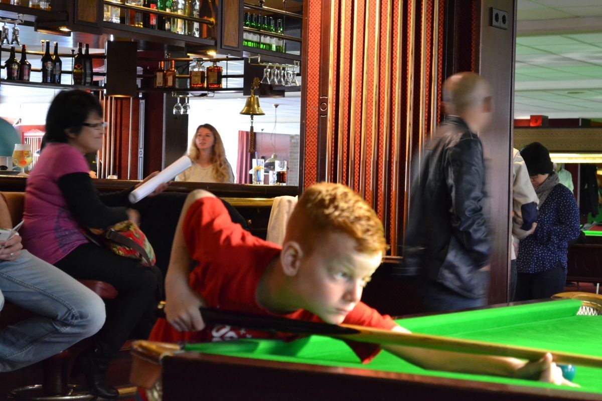 Jongen speelt pool in een bar, met een keu. Anderen zitten, blonde vrouw op de achtergrond bij de bar.