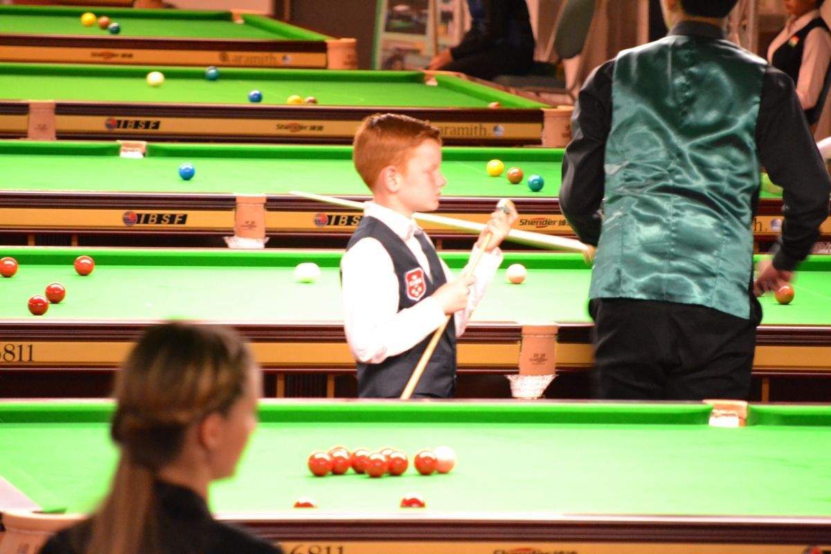 Een jonge roodharige jongen speelt snooker, met een keu in zijn hand. Meerdere snookertafels in een grote ruimte.