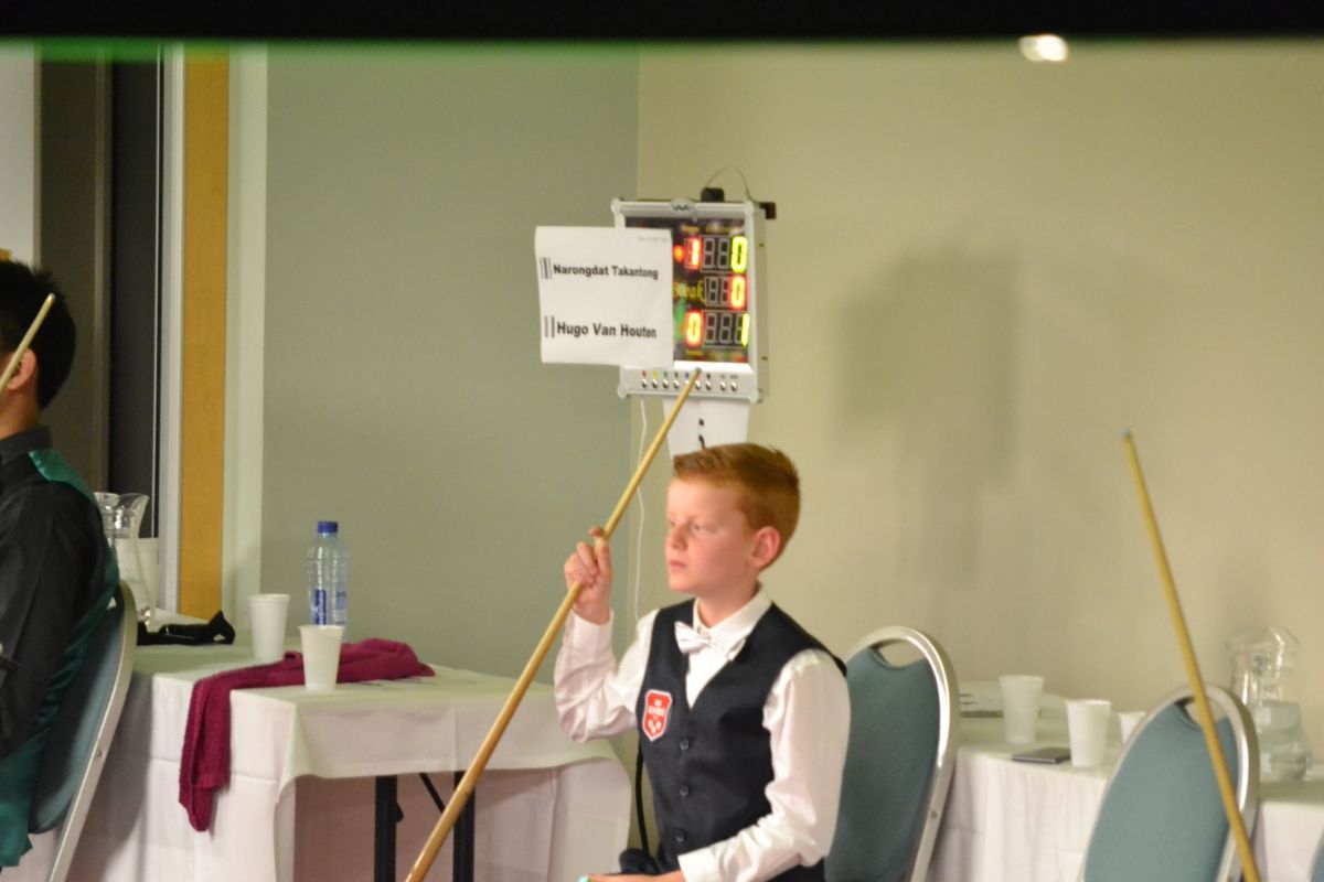 Een jongen in een vest houdt een keu vast en kijkt naar een scorebord tijdens een snookerwedstrijd.