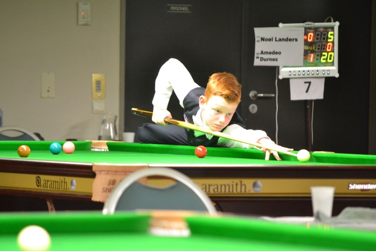 Een jonge jongen met rood haar speelt snooker en richt met zijn keu. Het scorebord toont een spel dat aan de gang is.