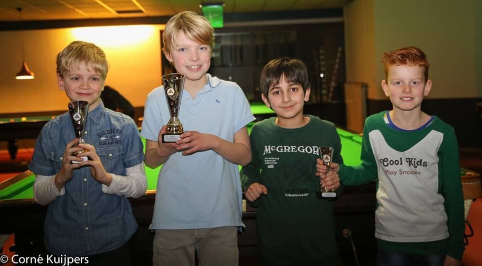 Vier lachende jongens met trofeeën in hun handen poseren in een snookerhal.