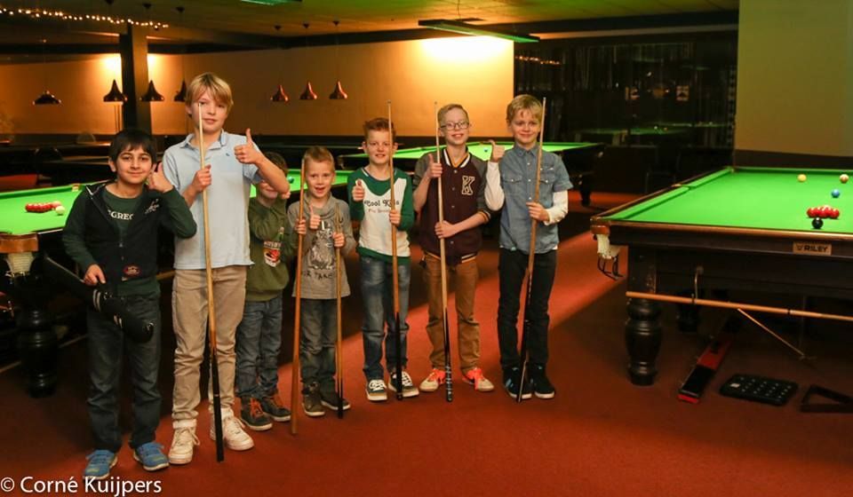Een groep jongens lacht en houdt keuen vast in een snookerhal.