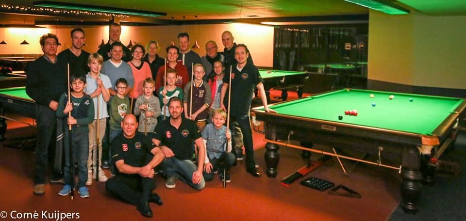 Een groep mensen, waaronder kinderen, poseert rond snookertafels in een biljartzaal.