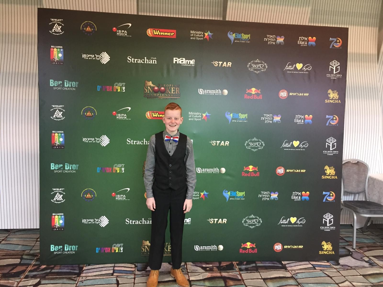 Een jonge jongen in formele kleding staat voor een groene achtergrond met bedrijfslogo's.