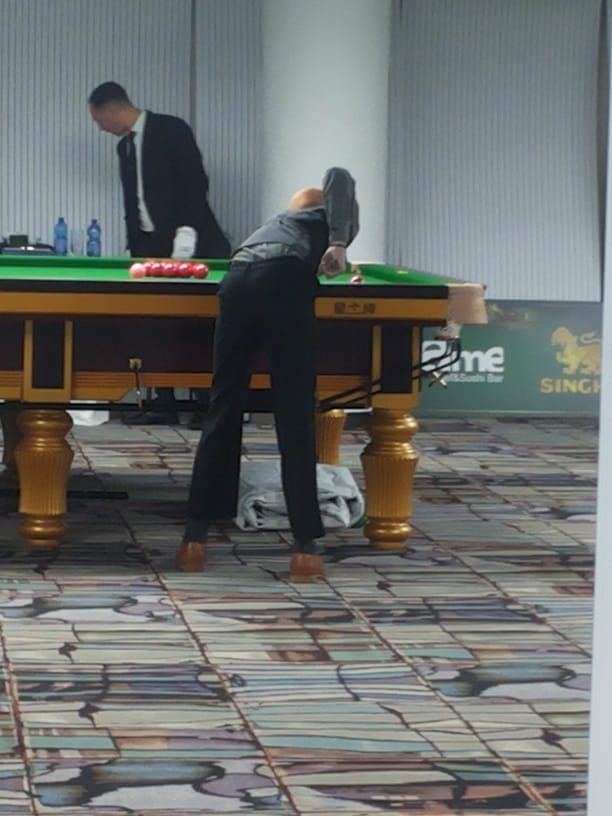 Een persoon die snooker speelt, hangt met een keu over een tafel; een ander kijkt toe. Binnen.