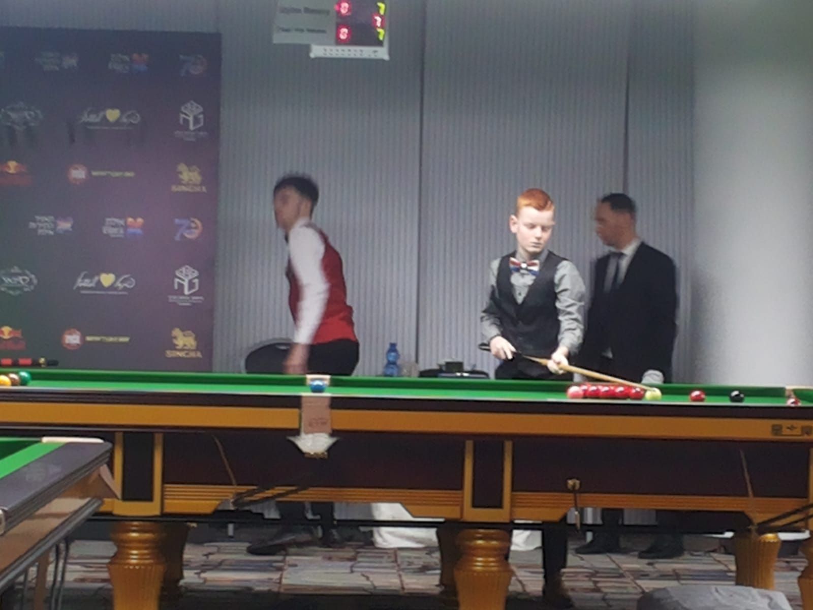 Snookerwedstrijd: Twee spelers en een scheidsrechter aan tafel. Een speler in een rood vest loopt, een ander mikt, de score wordt boven weergegeven.