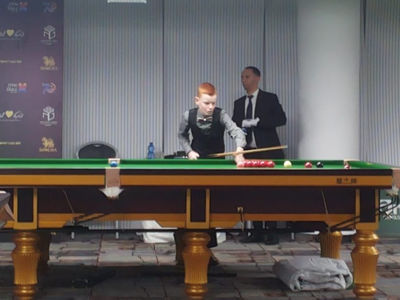 Een jongen met rood haar speelt snooker en richt een bal op een groene tafel. Een man in pak kijkt op de achtergrond toe.