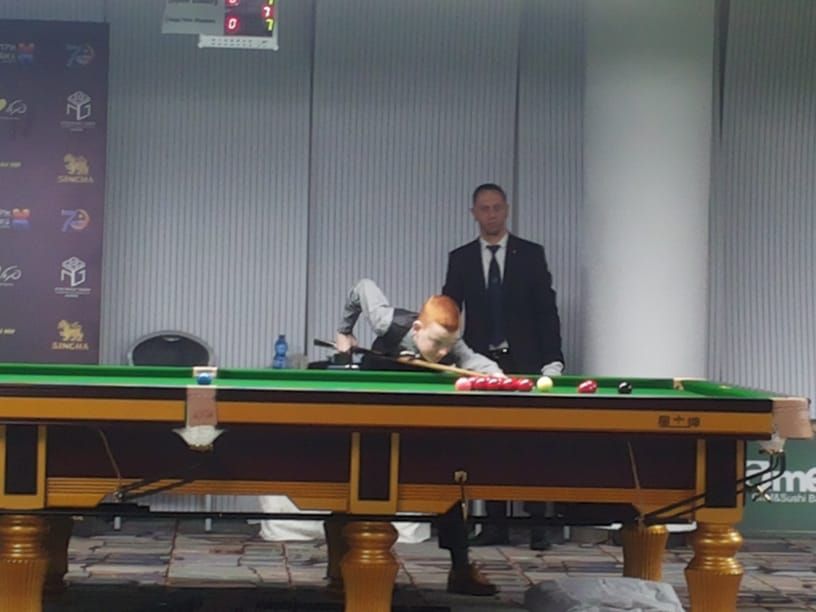 Een snookerspeler probeert een bal te scoren, terwijl de scheidsrechter op de achtergrond toekijkt.