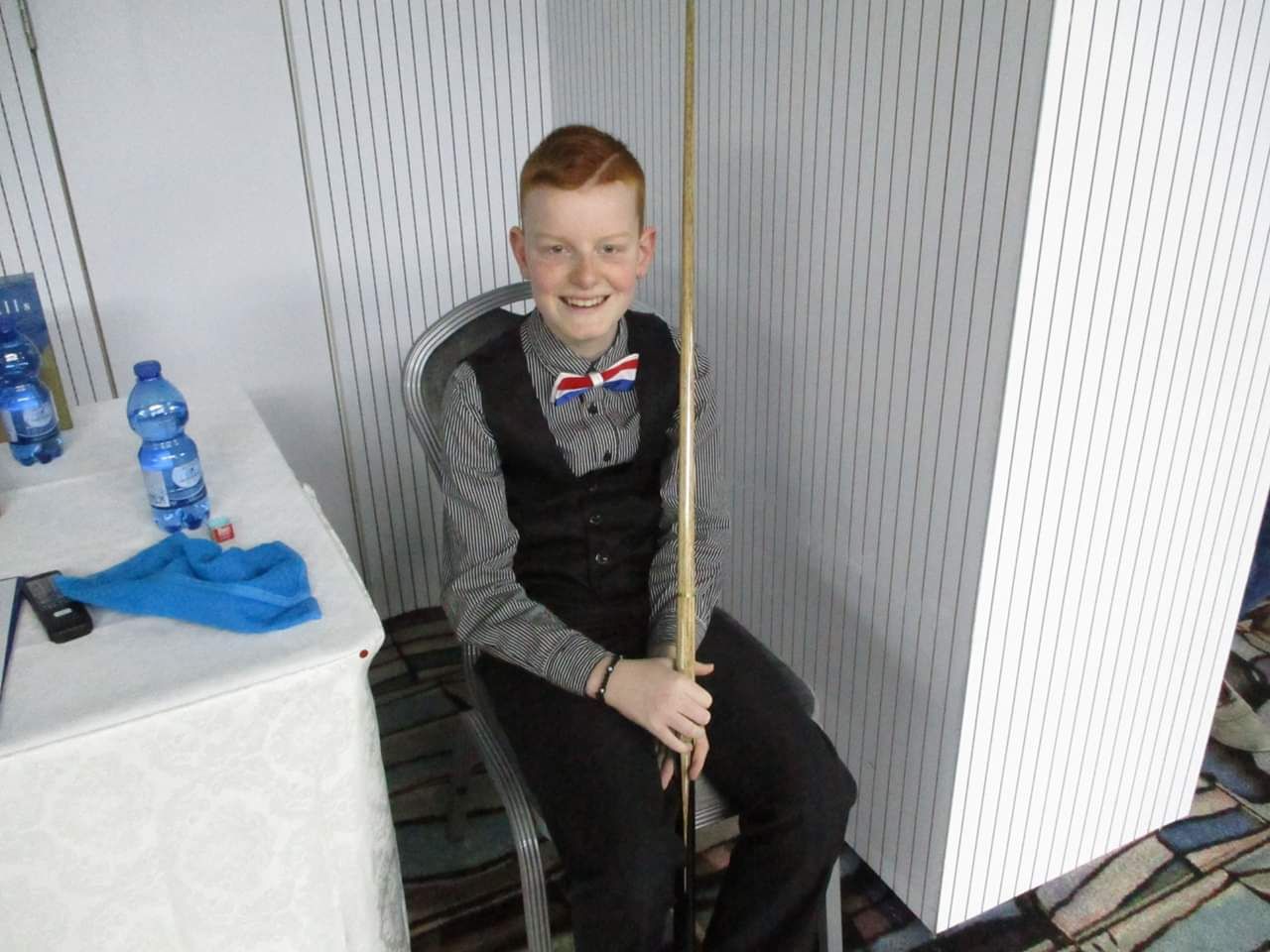 Een jonge roodharige jongen lacht, houdt een keu vast, draagt ​​een vlinderdasje en zit aan een tafel.