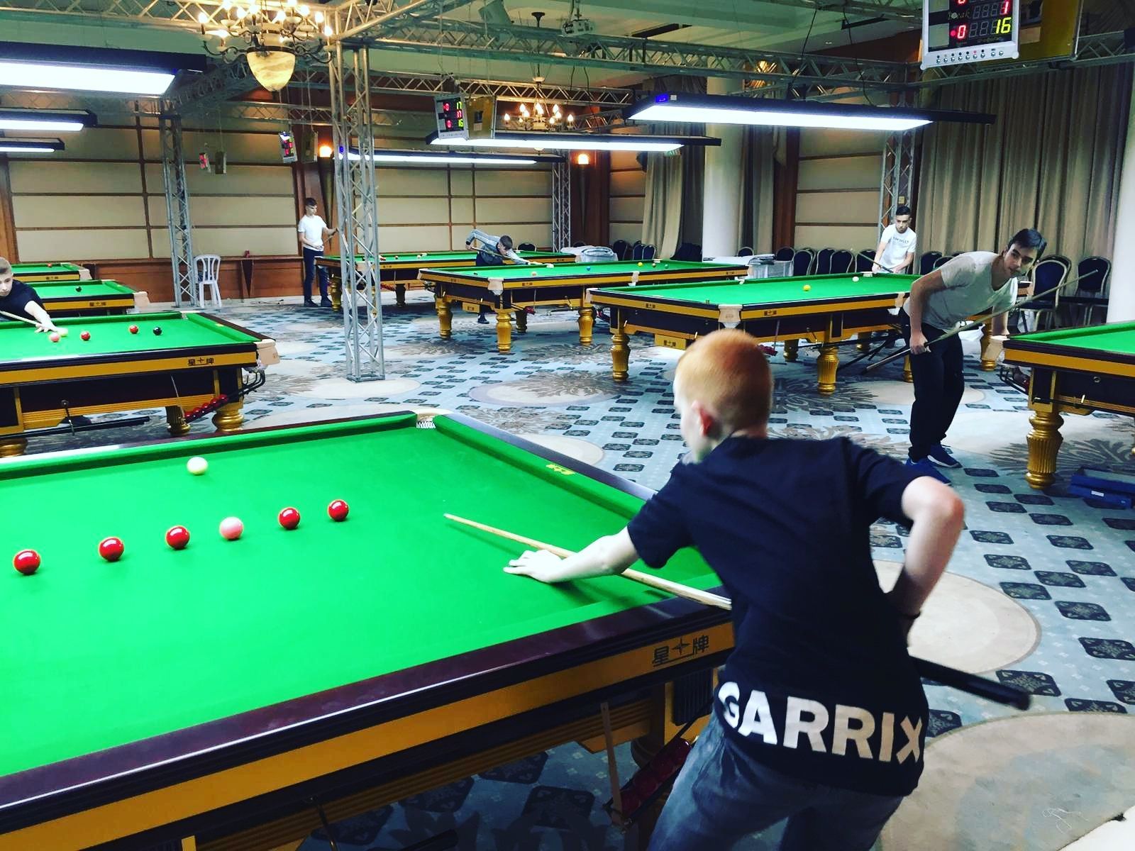 Een jongeman met rood haar bereidt zich voor op een snookertafel in een felverlichte snookerhal.
