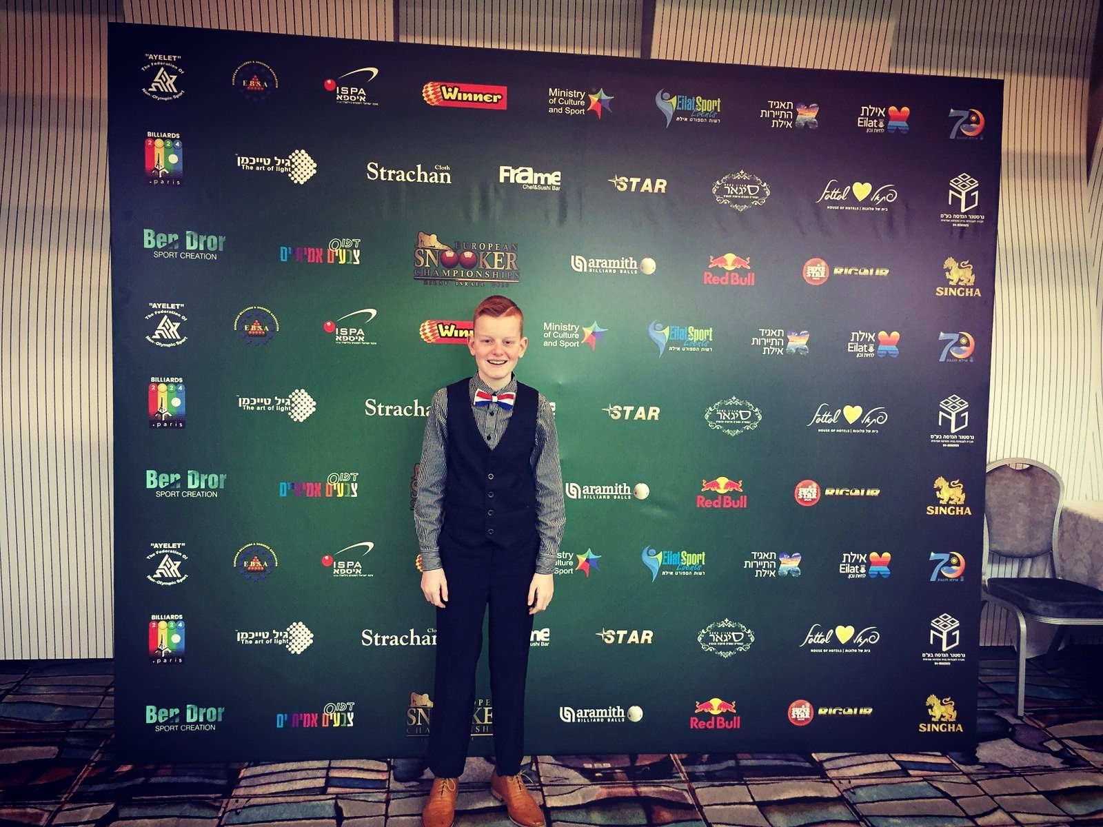 Een jongen in formele kleding staat voor een donkergroene achtergrond met logo's.